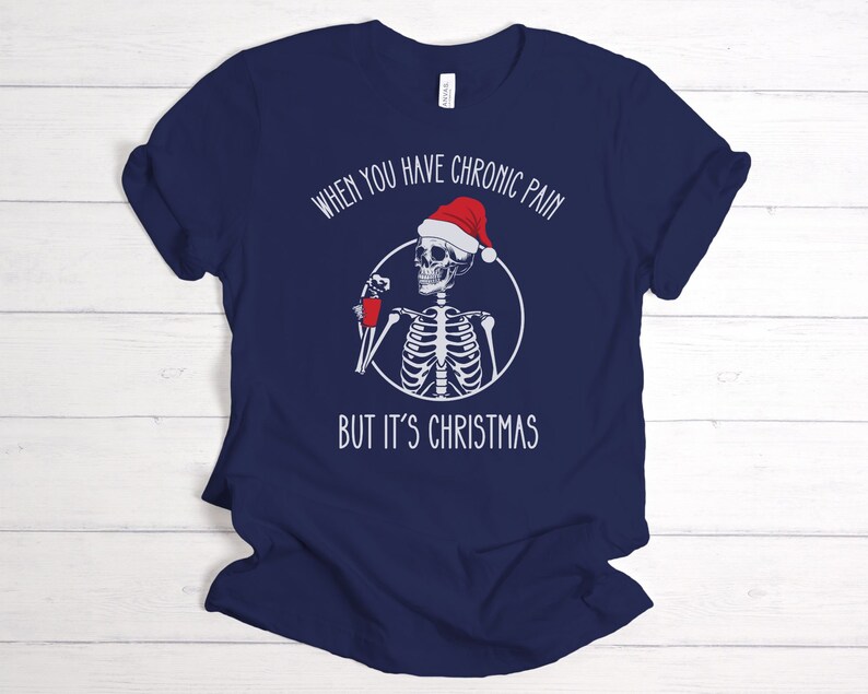 May include: Navy blue t-shirt with a white skeleton wearing a Santa hat and holding a red cup. The text on the shirt reads "When you have chronic pain but it's Christmas".