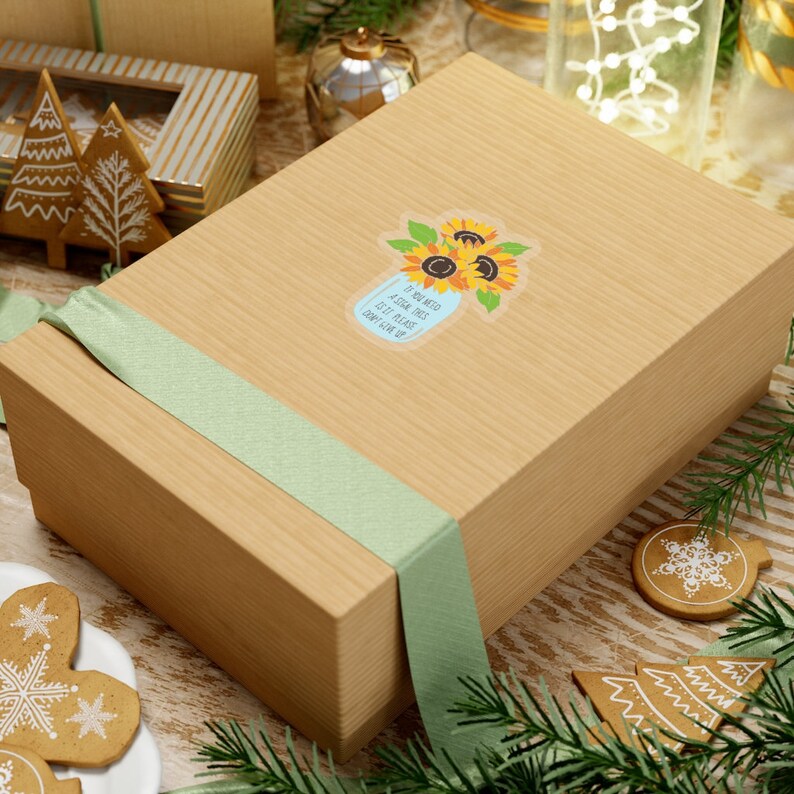 May include: A brown gift box with a green ribbon tied around it. The box has a sticker on it that says "If you need a sign, this is it. Please don't give up." The sticker features a blue mason jar with sunflowers.