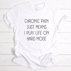 May include: White t-shirt with black text that reads "Chronic pain just means I play life on hard mode."