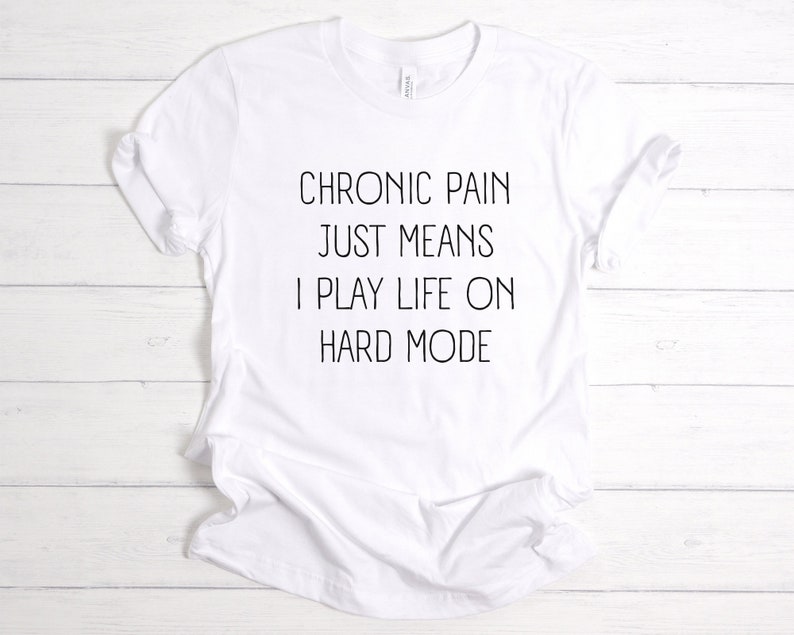 May include: White t-shirt with black text that reads "Chronic pain just means I play life on hard mode."