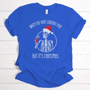 May include: A royal blue t-shirt with a white skeleton wearing a Santa hat and holding a red cup. The text on the shirt reads "When you have chronic pain but it's Christmas".