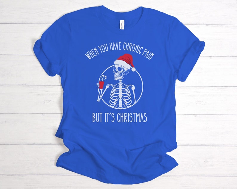 May include: A royal blue t-shirt with a white skeleton wearing a Santa hat and holding a red cup. The text on the shirt reads "When you have chronic pain but it's Christmas".