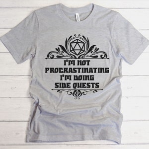 May include: A light gray t-shirt with the text "I'm not procrastinating I'm doing side quests" in black. The text is framed by decorative flourishes and a geometric shape. The shirt is made of a soft material.