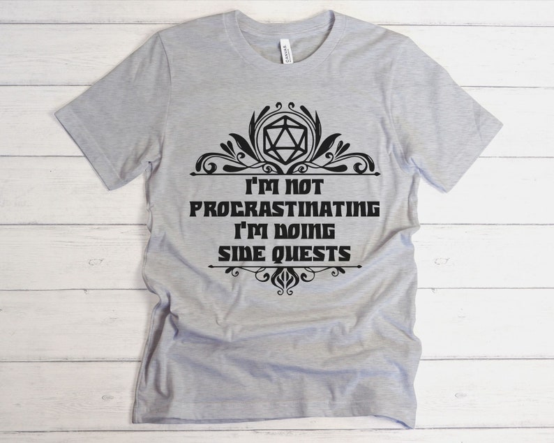May include: A light gray t-shirt with the text "I'm not procrastinating I'm doing side quests" in black. The text is framed by decorative flourishes and a geometric shape. The shirt is made of a soft material.