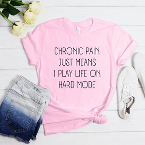 May include: Pink t-shirt with the text "Chronic pain just means I play life on hard mode" printed in black.