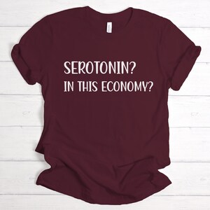 Pu&ograve; includere: Una maglietta color borgogna con testo bianco che dice "SEROTONIN? IN THIS ECONOMY?"