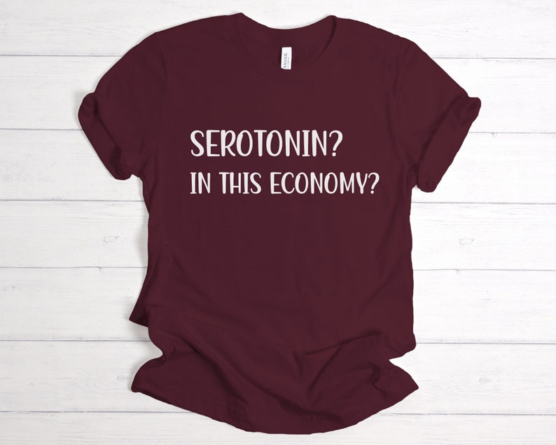 Pu&ograve; includere: Una maglietta color borgogna con testo bianco che dice "SEROTONIN? IN THIS ECONOMY?"