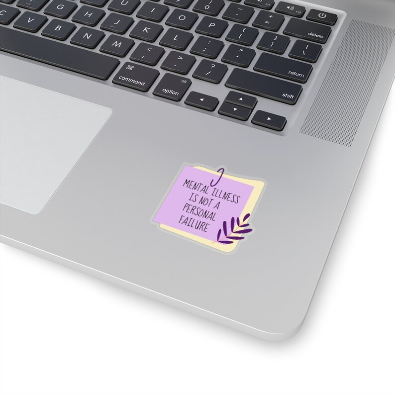 Puede incluir: Una pegatina con el texto "MENTAL ILLNESS IS NOT A PERSONAL FAILURE" en negro sobre una nota adhesiva morada, sujeta por un clip plateado. Un peque&ntilde;o dise&ntilde;o de hoja morada est&aacute; en la parte inferior derecha. La pegatina est&aacute; en un port&aacute;til.