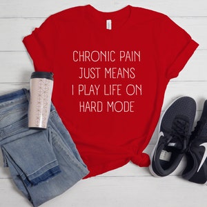 May include: Red t-shirt with white text that reads "Chronic pain just means I play life on hard mode". The shirt is laid out on a white wooden surface with a pair of black trainers, a pair of blue jeans, and a pink travel mug.