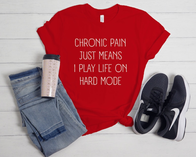May include: Red t-shirt with white text that reads "Chronic pain just means I play life on hard mode". The shirt is laid out on a white wooden surface with a pair of black trainers, a pair of blue jeans, and a pink travel mug.