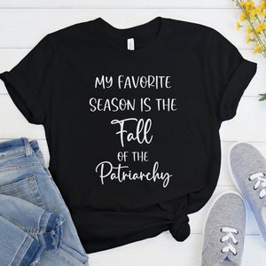 Op de afbeelding: Zwart T-shirt met witte tekst die luidt: "Mijn favoriete seizoen is de val van het patriarchaat."