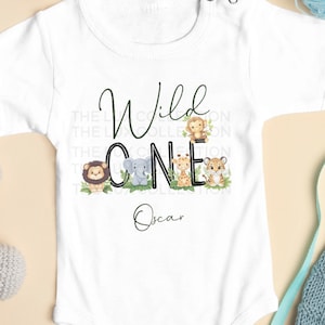 Puede incluir: Body de bebé blanco con el texto "Wild One" en escritura negra y verde, con ilustraciones de animales de dibujos animados. El nombre "Oscar" está impreso debajo. El body tiene mangas cortas y cierres de broche.
