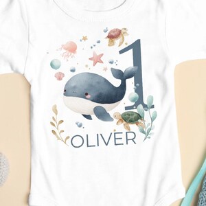 Op de afbeelding: Witte babybody met een walvis en het cijfer 1, omringd door zeedieren. De naam "OLIVER" staat eronder gedrukt. Een blauwe gebreide romper, een grijze nijlpaardknuffel en andere babyartikelen zijn eromheen geplaatst.