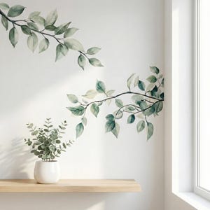 Op de afbeelding: Muurstickers met bladerrijke takken in aquarel-stijl in groen- en grijstinten. Een kleine potplant met groene bladeren staat op een houten plank. De muur is wit en rechts is een raam zichtbaar.