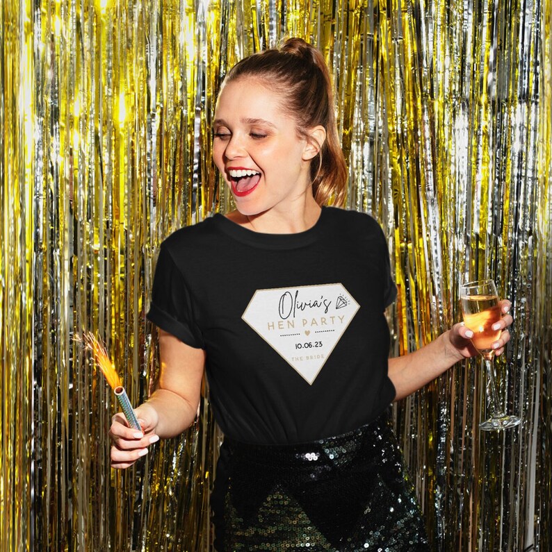 May include: Black t-shirt with a diamond-shaped graphic that reads "Olivia's Hen Party" with a date. The woman in the image is holding a sparkler and a glass of champagne. The background is a gold metallic curtain.