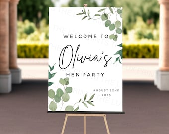 May include: A white sign on a wooden easel with the text "WELCOME TO Olivia's HEN PARTY AUGUST 22ND 2023". The sign is decorated with green eucalyptus leaves and branches.