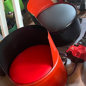 Puede incluir: Dos sillas de barril recicladas, una con asiento de cuero negro y la otra con asiento de cuero rojo. Ambas sillas están hechas de barriles de metal y tienen un aspecto industrial único.