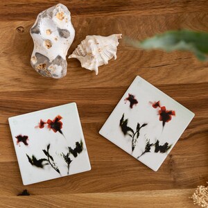 Op de afbeelding: Twee witte vierkante onderzetters met zwarte en rode bloemenmotieven. De onderzetters liggen op een houten oppervlak met een schelp en een steen.