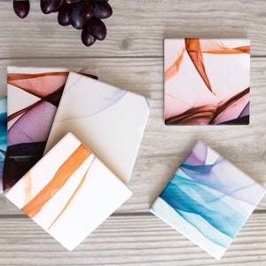 Può includere: Una collezione di sottobicchieri quadrati in ceramica con disegni astratti nei toni del blu, viola e arancione. I sottobicchieri sono disposti su una superficie di legno chiaro, con un grappolo d'uva nell'angolo in alto a sinistra.