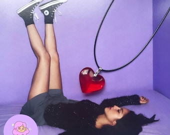 May include: A red heart-shaped pendant necklace with a black cord. The pendant is suspended against a purple background. The image also features black Converse sneakers and the text "VELVET ACCESSORIES".