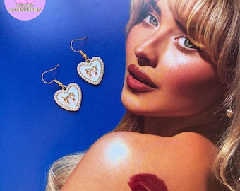 May include: A pair of gold heart-shaped earrings with a white enamel finish and a gold bow design. The earrings have a pearl-like border.