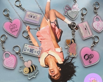 May include: A collection of acrylic keychains featuring various designs including a pink heart with text, a purple cassette tape, a California license plate, a bee, a padlock, and a figure holding a heart, all attached to silver metal lobster clasps.