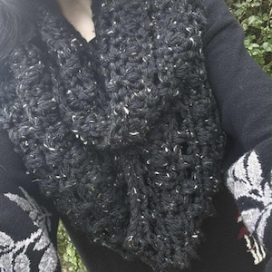 Puede incluir: Bufanda infinita de punto grueso gris oscuro con motas blancas. La bufanda es gruesa y parece cálida, adecuada para el clima frío. La imagen también muestra un suéter negro y una manga con un diseño floral.