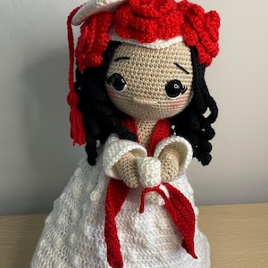 May include: A handmade crochet doll in a graduation outfit. The doll features black hair, a tan face, and a white gown with red details. The graduation cap is red and white, and the doll is approximately 20 cm tall.