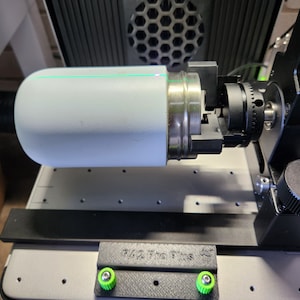 May include: A white cylindrical object is held in a black clamp with a green laser line across the centre. The object is attached to a black metal arm with a black knob. The arm is attached to a black base with a green knob. The base is on a grey surface with a black label that reads "R/2 Pro Plus".