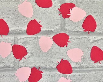 May include: A collection of pink and red paper cutouts of strawberries. The cutouts are scattered on a white brick background.