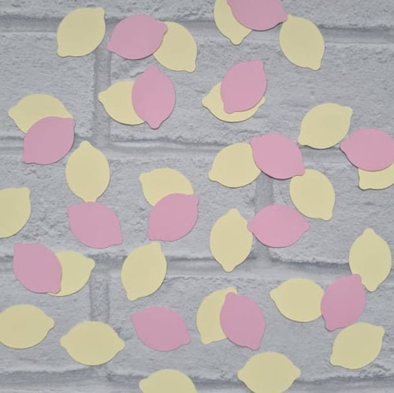 May include: A collection of yellow and pink paper lemon cutouts scattered on a white brick background.