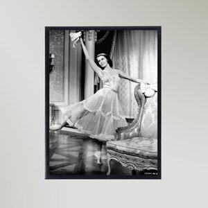 Könnte beinhalten: Ein Schwarzweißfoto einer Frau in einem weißen Tüllkleid, die in einem Raum mit einem Stuhl steht. Sie hält einen weißen Federboa in ihrer rechten Hand und schaut nach links. Der Raum hat einen gemusterten Teppich und ein großes Fenster.