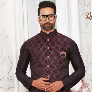 Può includere: Una kurta nera con un gilet a motivi rosa e neri. Il gilet ha una catena dorata e un ornamento decorativo. L'uomo indossa pantaloni neri e scarpe marroni.