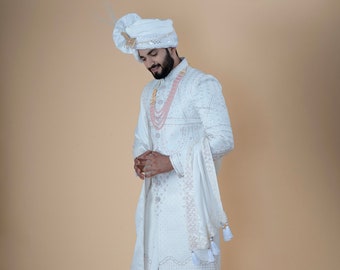 May include: A white embroidered sherwani suit with matching trousers and shoes. The outfit includes a turban, a scarf, and a pink beaded necklace. The traditional Indian formal wear is suitable for weddings and special occasions.