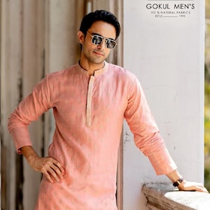 May include: A light peach-colored kurta with gold detailing. The long-sleeved garment has a mandarin collar and is made of pure linen silk. The image includes text that reads "GOKUL MEN'S 100% NATURAL FABRICS EST.D 1993" and "Kurta - Pure Lilen Silk".