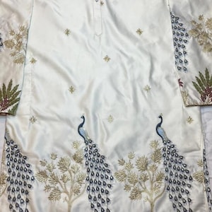 May include: A long-sleeved white tunic featuring a button-down placket at the neckline. The fabric is decorated with embroidered gold star motifs and a detailed lower panel showcasing two blue peacocks, golden trees, and small brown deer figures.
