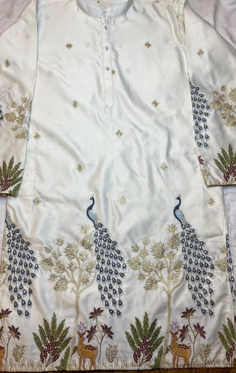 May include: A long-sleeved white tunic featuring a button-down placket at the neckline. The fabric is decorated with embroidered gold star motifs and a detailed lower panel showcasing two blue peacocks, golden trees, and small brown deer figures.