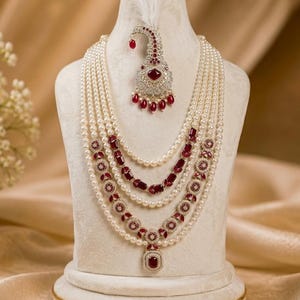 May include: A multi-strand pearl necklace featuring deep red gemstone accents and intricate silver-toned metalwork. The set includes a matching teardrop-shaped headpiece adorned with a white feather, red stones, and clear crystals on a display bust.