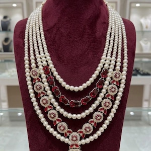 May include: A multi-strand pearl necklace with ruby and diamond accents. The necklace features five strands of white pearls, interspersed with ruby and diamond clusters and a large ruby pendant. The necklace is displayed on a burgundy velvet bust.