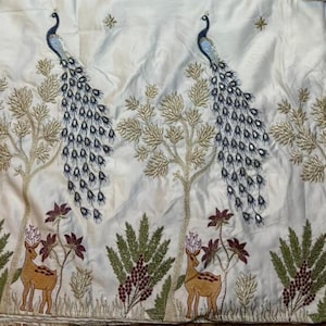 May include: A white fabric featuring an intricate embroidered pattern of two blue and gold peacocks perched in trees, with two brown deer standing below among green foliage and small maroon flowers, accented with small gold star motifs.