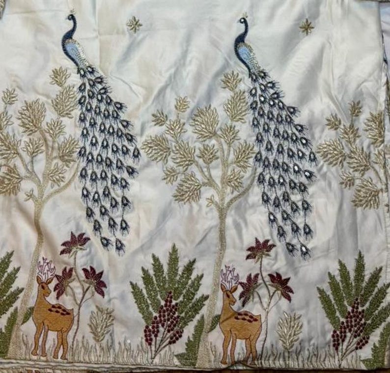 May include: A white fabric featuring an intricate embroidered pattern of two blue and gold peacocks perched in trees, with two brown deer standing below among green foliage and small maroon flowers, accented with small gold star motifs.