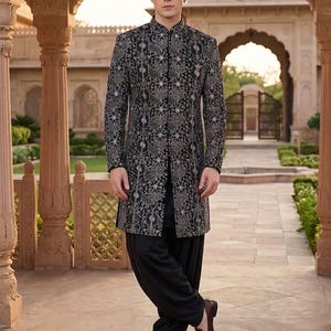 May include: A black sherwani featuring a detailed silver and grey geometric pattern with long sleeves and a high collar, paired with solid black trousers and brown leather loafers. The garment has a structured fit and reaches mid-thigh length.