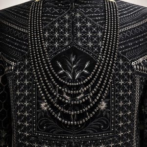 Könnte beinhalten: Nahaufnahme einer schwarzen Jacke mit aufwendigen Stickereien und Perlenarbeiten. Die Jacke hat einen hohen Kragen und verzierte Muster, darunter florale und geometrische Designs. Silberne Akzente unterstreichen die Details.
