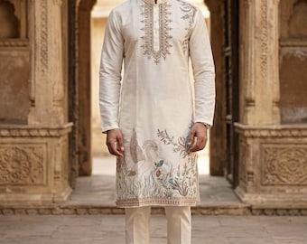 May include: Off-white kurta pajama set with intricate embroidery and a nature-inspired design. The long-sleeved kurta features detailed patterns around the collar and a decorative motif with a bird and foliage. The matching pants complete the ensemble.
