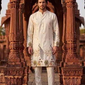 May include: An off-white long-sleeved kurta featuring a mandarin collar and a button-down placket. The garment is decorated with embroidered peacock and deer motifs along the hemline and sleeves, paired with matching off-white trousers and brown loafers.