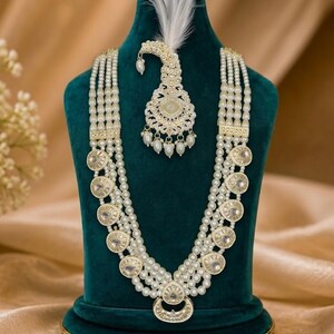 Peut inclure: Un ensemble collier multi-rangs et bijou de tête composé de perles blanches et d'accents métalliques circulaires avec des pierres transparentes. Le collier comporte cinq rangs de perles, tandis que le bijou de tête inclut une plume blanche.