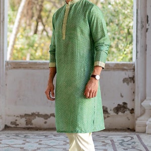 Puede incluir: Una camisa verde de manga larga con un dise&ntilde;o estampado blanco y dorado. La camisa tiene un cuello mandar&iacute;n y cierre de bot&oacute;n. El modelo lleva pantalones blancos y zapatos blancos.