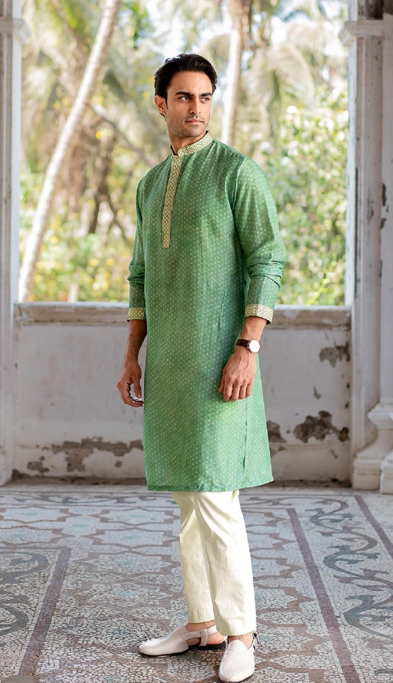 Puede incluir: Una camisa verde de manga larga con un dise&ntilde;o estampado blanco y dorado. La camisa tiene un cuello mandar&iacute;n y cierre de bot&oacute;n. El modelo lleva pantalones blancos y zapatos blancos.