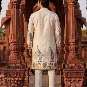 May include: An off-white long-sleeved tunic featuring intricate embroidery of blue peacocks, golden trees, and small deer along the hem and sleeves, paired with matching off-white trousers. The garment has a mandarin collar and a buttoned front.
