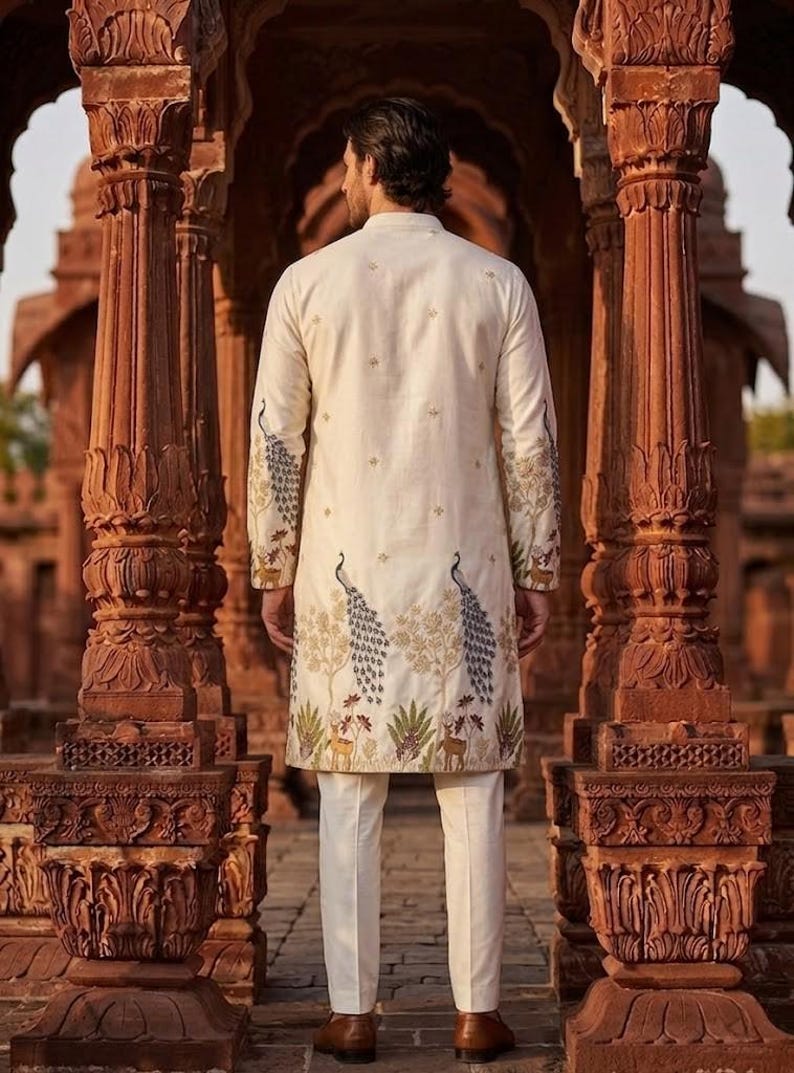 May include: An off-white long-sleeved tunic featuring intricate embroidery of blue peacocks, golden trees, and small deer along the hem and sleeves, paired with matching off-white trousers. The garment has a mandarin collar and a buttoned front.